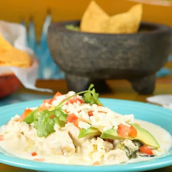 Creamy Mexican chicken dish topped with avocado, tomatoes, cheese, and cilantro, with guacamole in background.